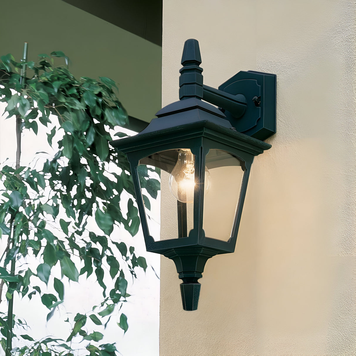 The Chapel 1 Light Outdoor Mini Down Wall Lantern in black, with its glowing bulb, is elegantly mounted on a beige wall as part of sophisticated die-cast aluminum outdoor lighting. Nearby, a leafy plant casts shadows across the lantern's classic hexagonal glass enclosure.