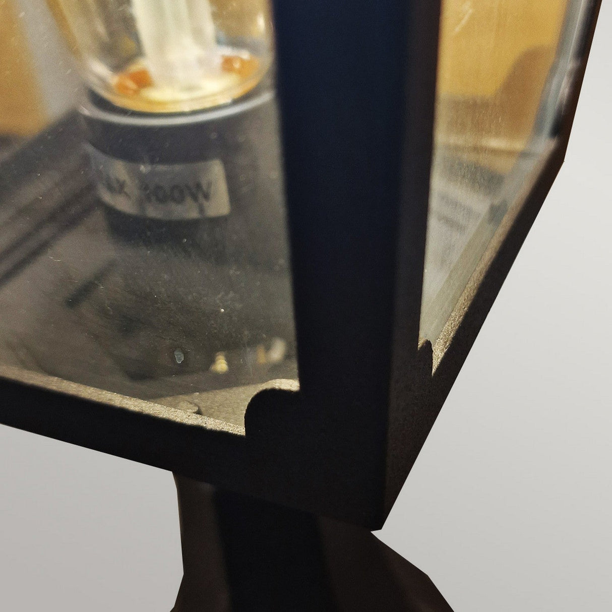 Close-up of the Chapel 1 Light Outdoor Mini Pedestal Lantern in black, showcasing its dark-colored, die-cast aluminum frame. Inside, a visible light bulb with a MAX 100W label is featured. The IP44-rated lantern creates a beautiful soft focus effect as it reflects light.