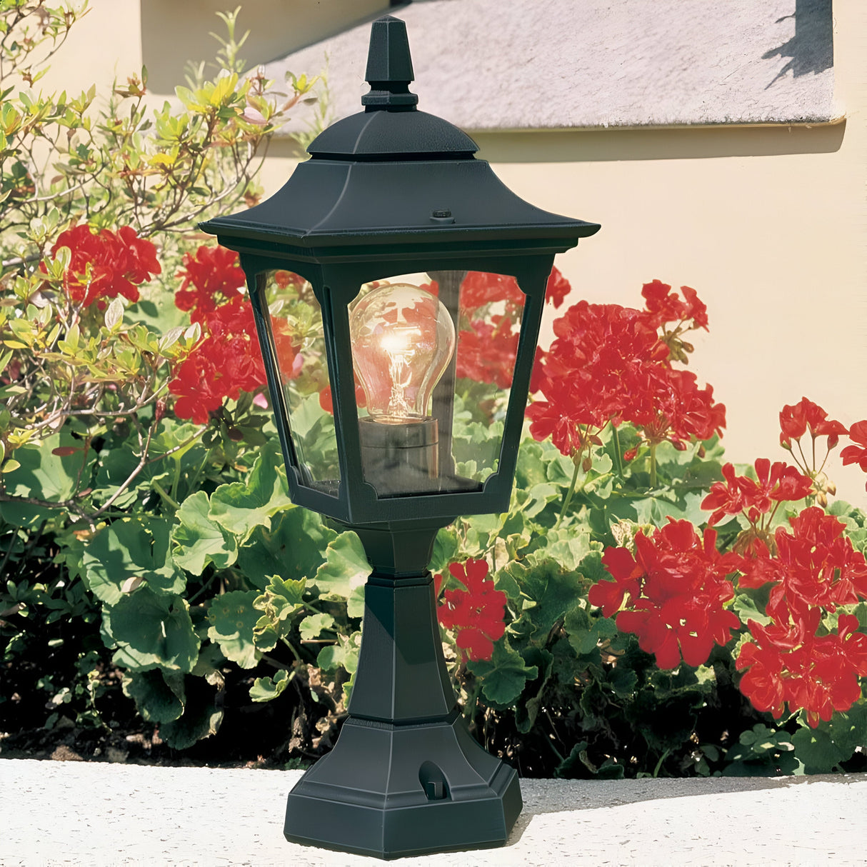 A Chapel 1 Light Outdoor Mini Pedestal Lantern in black die-cast aluminum sits on a white ledge surrounded by vibrant red geraniums and lush green leaves. The IP44-rated lantern is turned on, emitting a warm glow against a beige wall with a small stone accent.