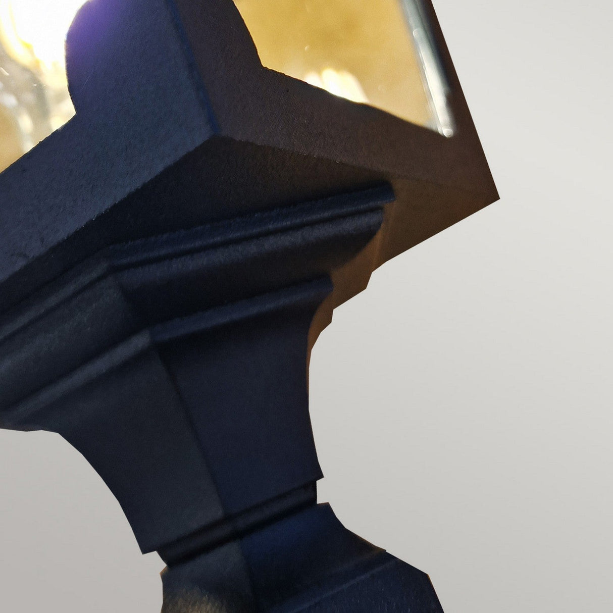 Close-up of the Chapel 1 Light Mini Pillar Lantern - Black, featuring a square glass enclosure and glowing brightly. Crafted from die-cast aluminium, this lantern is mounted on an ornate metal post against a plain grey background, showcasing the charm of classic outdoor lighting.