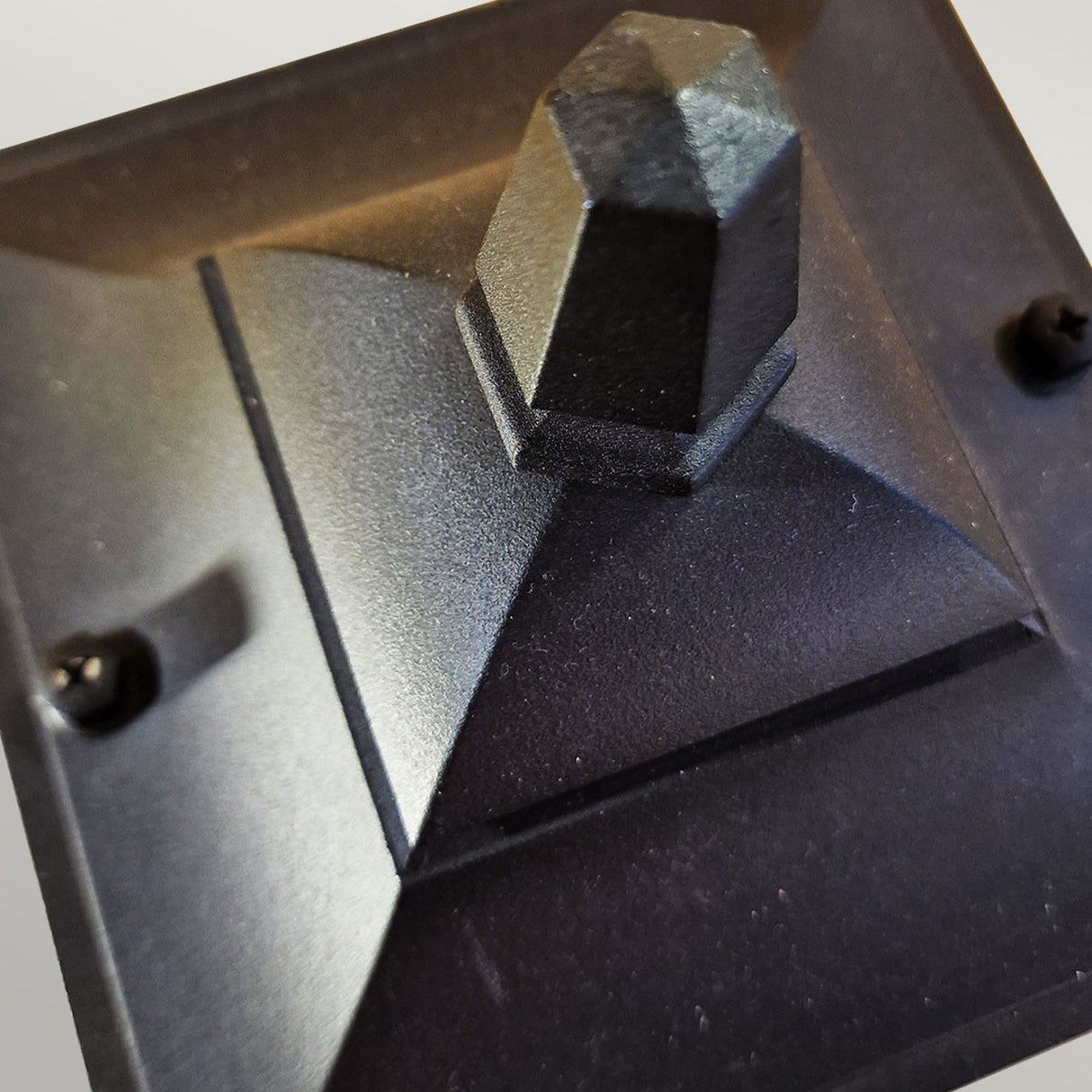 A close-up showcases a textured, black pyramid-shaped metal post cap made from die-cast aluminum, featuring a smooth, pointed top. Two visible screw heads are on either side of the cap to secure it to a surface. The blurred background draws attention to its geometric design, reminiscent of the Chapel 1 Light Mini Pillar Lantern - Black.