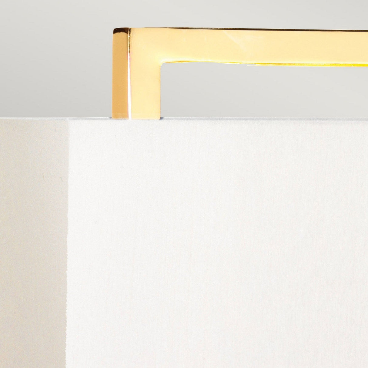 A close-up showcases the minimalistic design of the Cross Table Lamp with Ivory Shade, highlighting its polished gold handle against a white and beige textured surface. The diagonal positioning of the handle enhances the modern and elegant aesthetic against the ivory shade background.