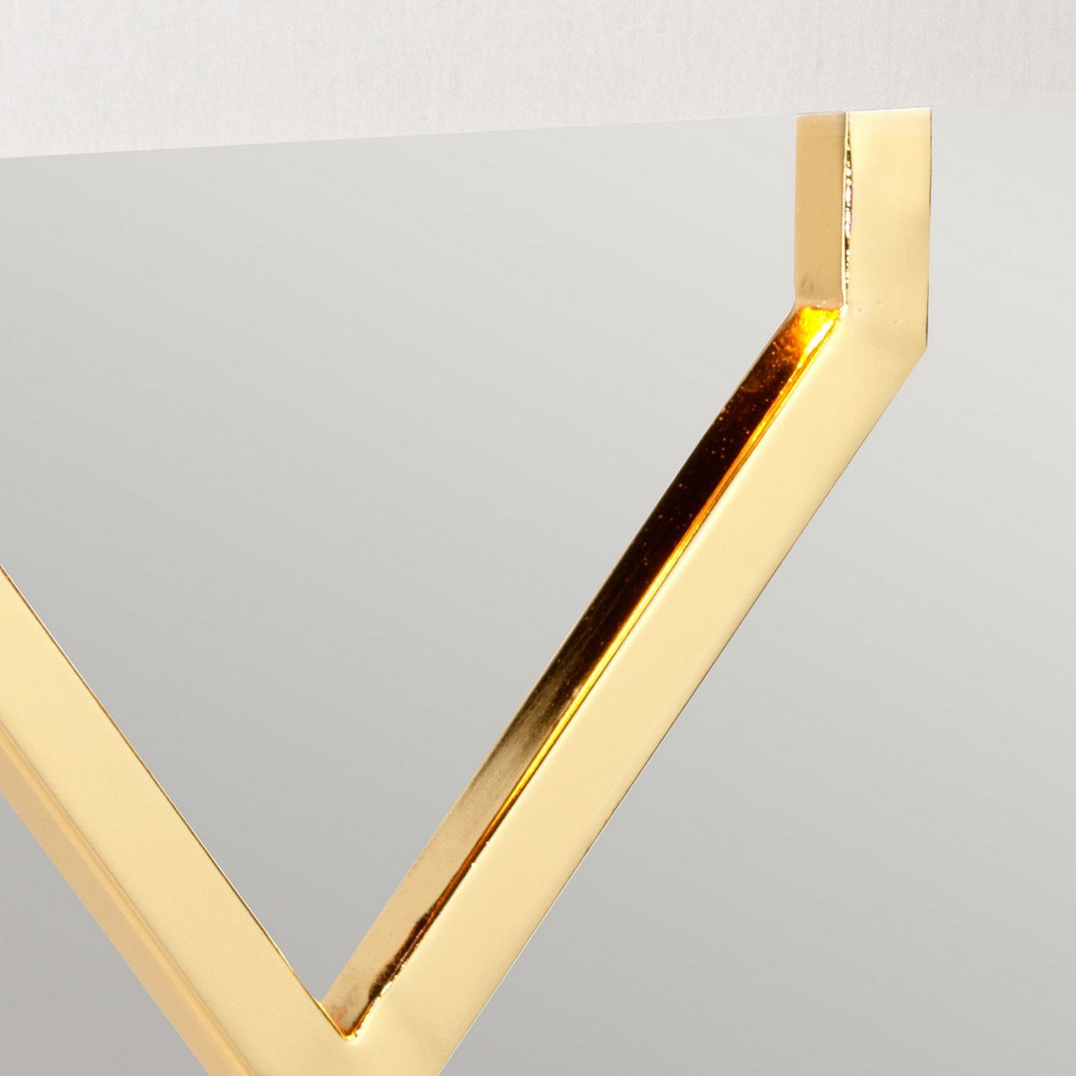 Close-up of a corner of a modern mirror with a frame reminiscent of the polished gold finish found in the Cross Table Lamp with Ivory Shade. The sleek, angular design and shiny metallic surface reflect light beautifully, creating an elegant contrast against the smooth mirror surface and complementing the ivory shade tones against the soft gray background.