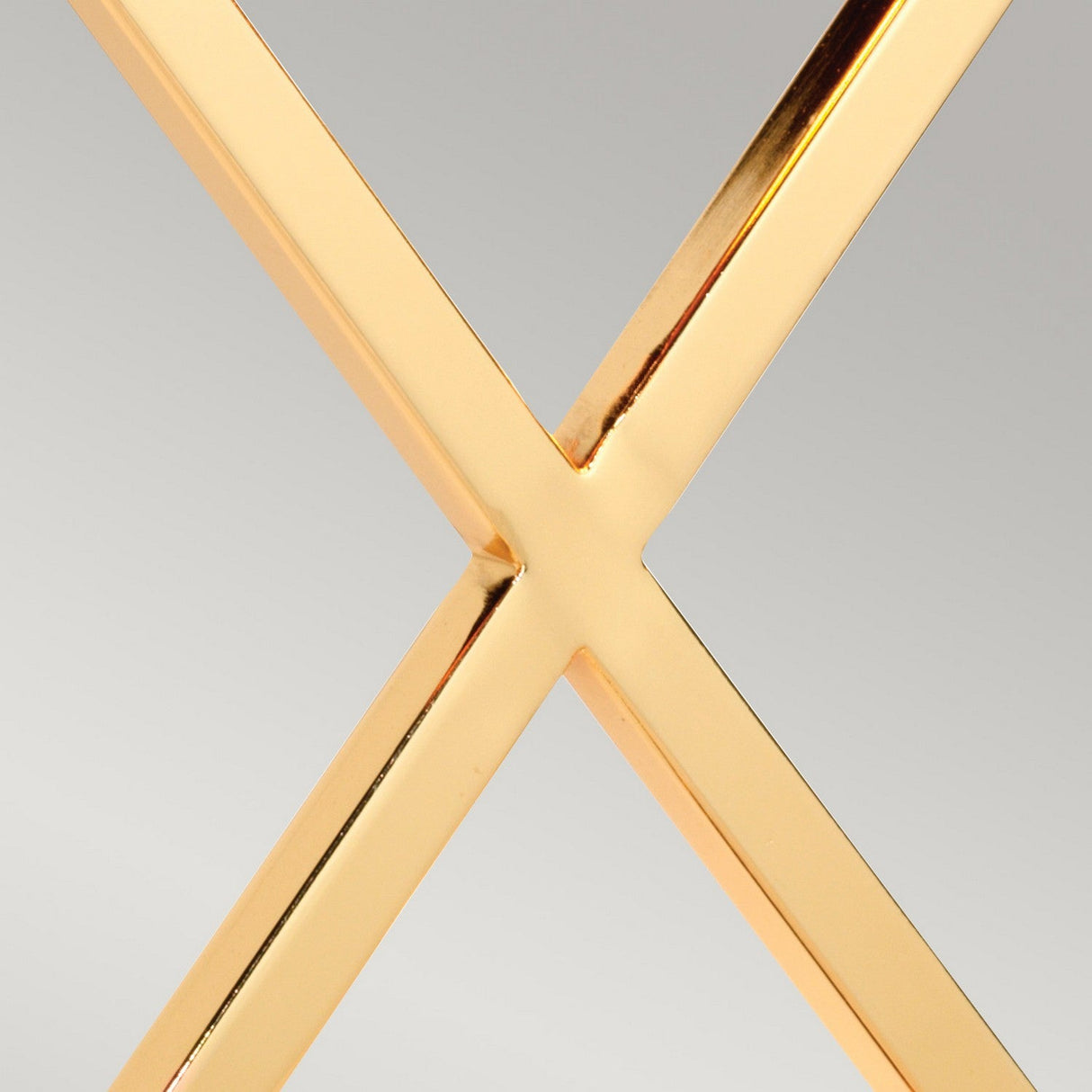 A close-up of a shiny gold metallic X shape against a neutral background resembles the polished gold base of the Cross Table Lamp with Ivory Shade. The intersecting lines have a smooth, polished surface that subtly reflects light, much like how the ivory shade softly illuminates the room.