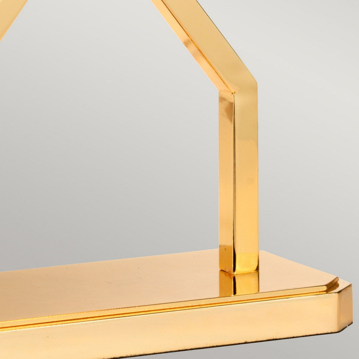 Close-up of the Cross Table Lamp With Blue Shade - Polished Gold, showcasing a polished gold base with a geometrically shaped, arch-like structure. Its smooth, reflective surface contrasts beautifully against the plain gray background, enhancing an elegant and modern ambiance.