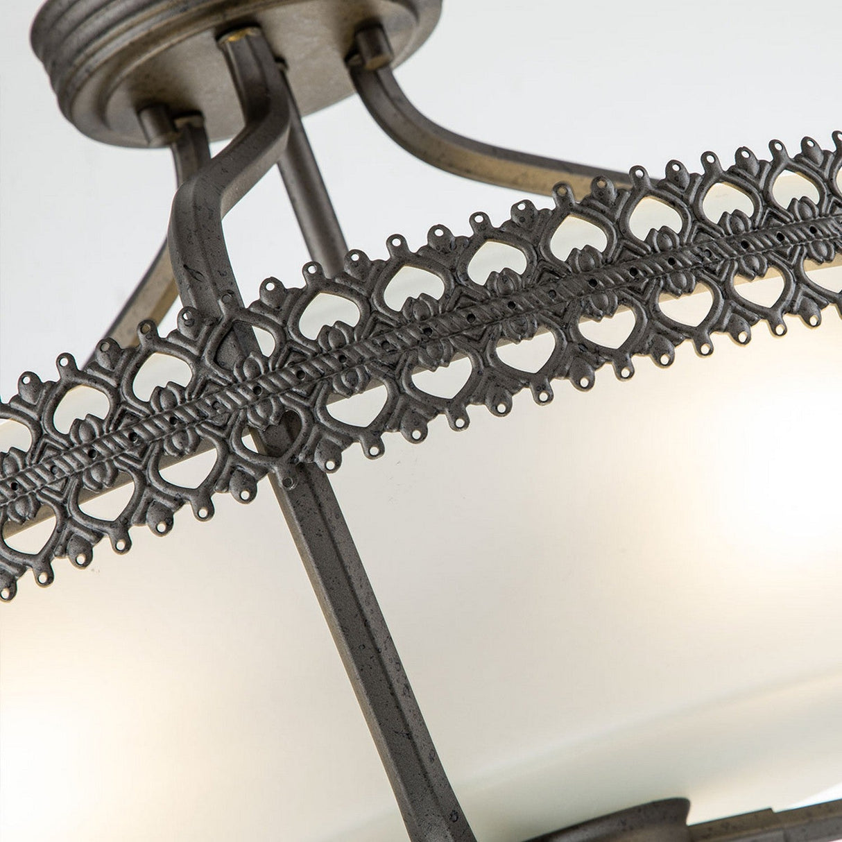 A close-up of the Crown 3 Light Semi Flush in graphite reveals its ornate design, featuring intricate lace-like detailing on the circular band. The light emits a gentle glow, highlighting the exquisite craftsmanship and architectural presence of this chandelier against a plain backdrop.