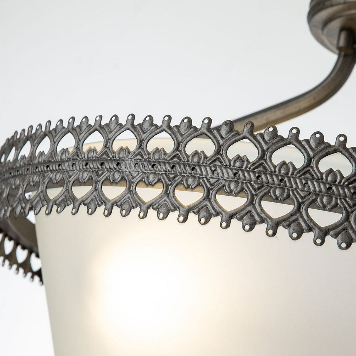 A close-up of the Crown 3 Light Semi Flush - Graphite lighting fixture showcases its intricate metal lace-like detailing around the edge. The metal is expertly interwoven, forming a decorative pattern reminiscent of architectural elegance, and it emits a soft glow beneath the metalwork.