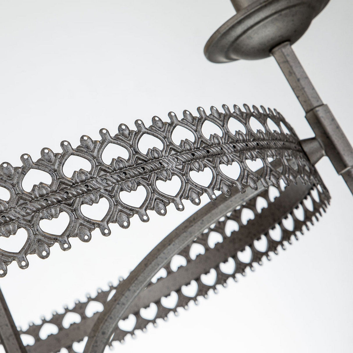 A close-up view of the Crown 3 Light Chandelier - Graphite showcases its ornate metal structure, adorned with intricate heart-shaped patterns that evoke a traditional influence. The finely detailed design features a repeating motif and a textured, graphite-like metallic finish set against a plain background.