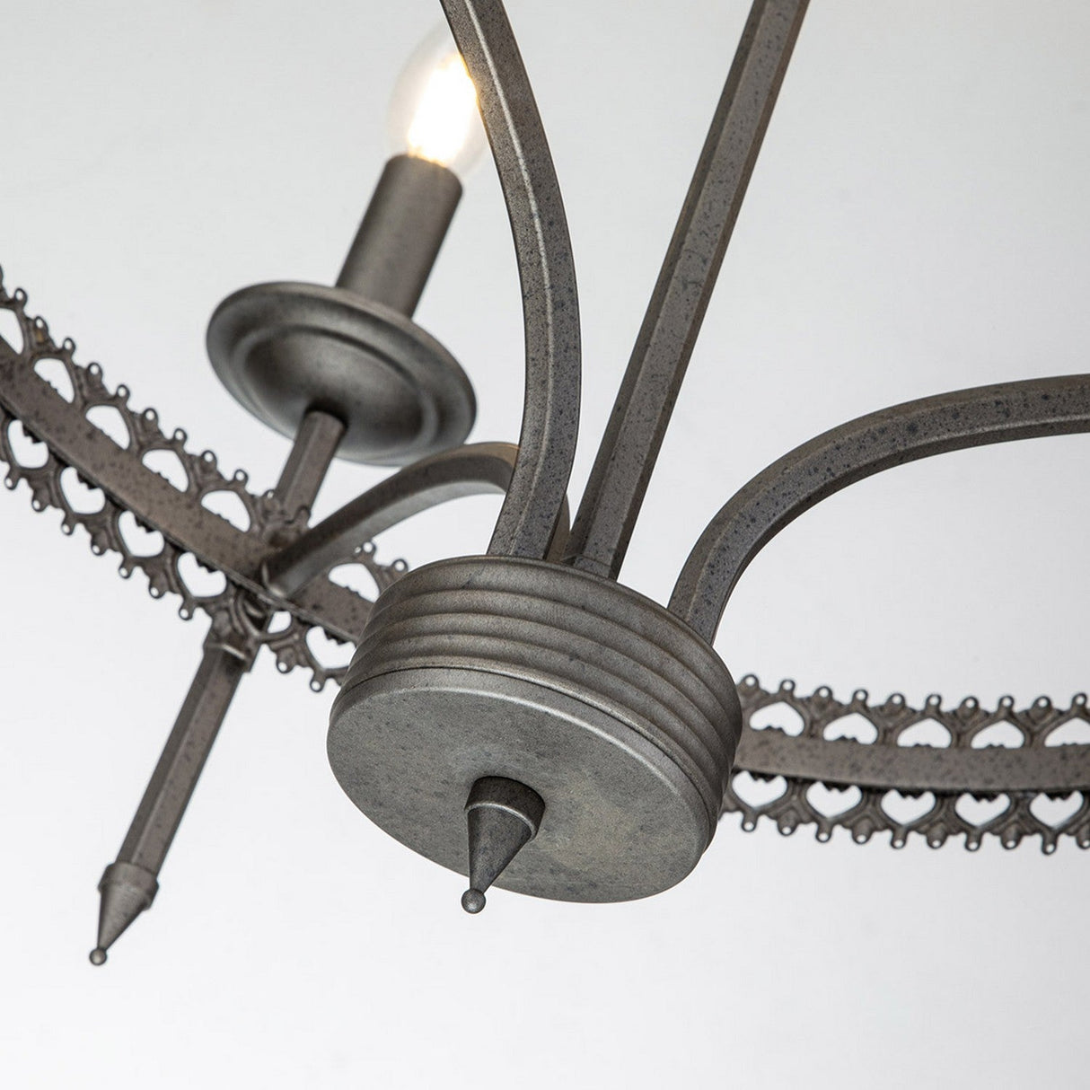 Up-close perspective of the Crown 3 Light Chandelier - Graphite, showcasing its traditional rustic metal design with a single exposed bulb. The chandelier features ornate curved metal arms and a decorative circular element, all adorned in a weathered, graphite-like industrial finish.
