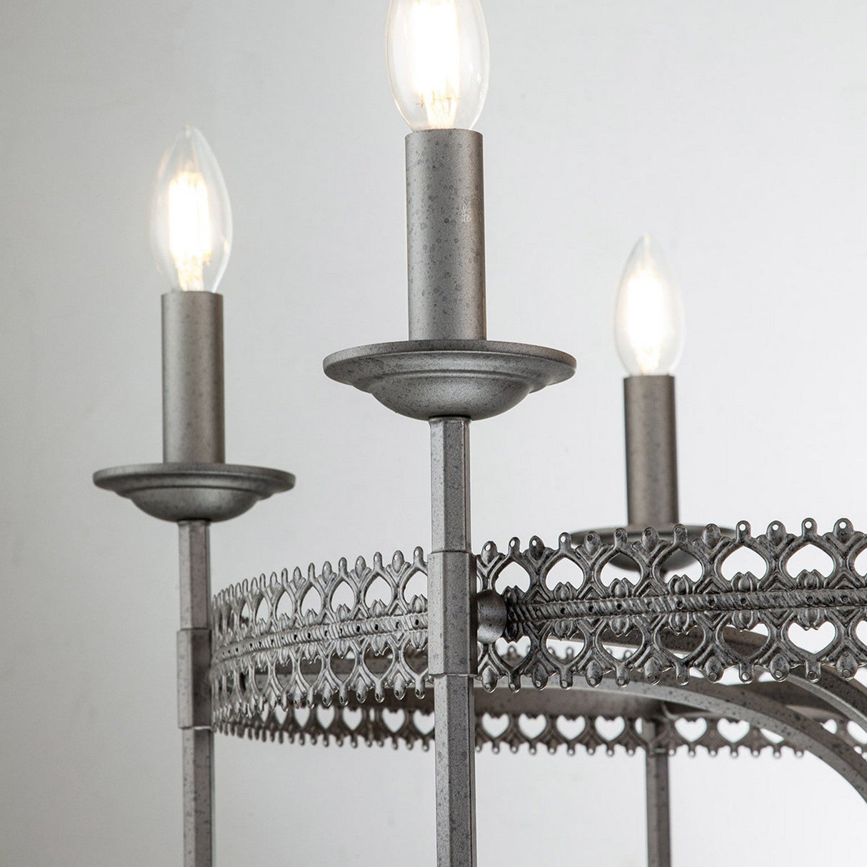 Close-up of the Crown 6 Light Chandelier - Graphite, showcasing its ornate metalwork and candle-style light fixtures. The bulbs are lit, casting a warm glow. This graphite chandelier features rustic gray metal with intricate patterns, exuding regal elegance in every detail.