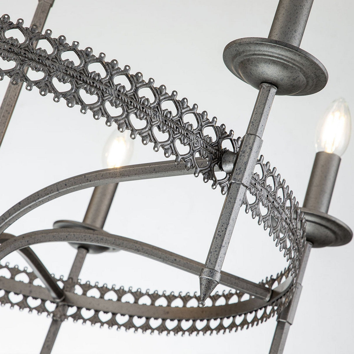 Close-up of the Crown 6 Light Chandelier - Graphite, an ornate metal masterpiece exuding regal elegance with intricate, lace-like patterns. It features candle-shaped bulbs and a rustic, antique graphite finish against a plain, light-colored wall.