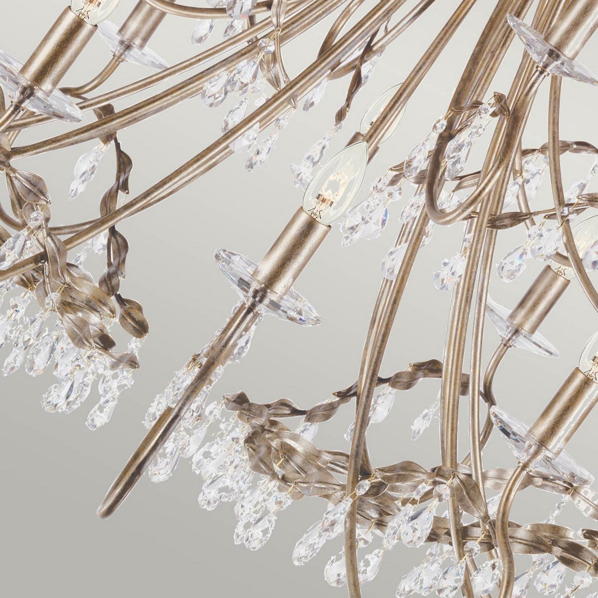 A close-up of the Christina 18 Light Chandelier in a silver and gold patina reveals its intricate metalwork and dangling crystal drops, along with candle-shaped light bulbs. The neutral-toned background accentuates the chandelier's elegant design.
