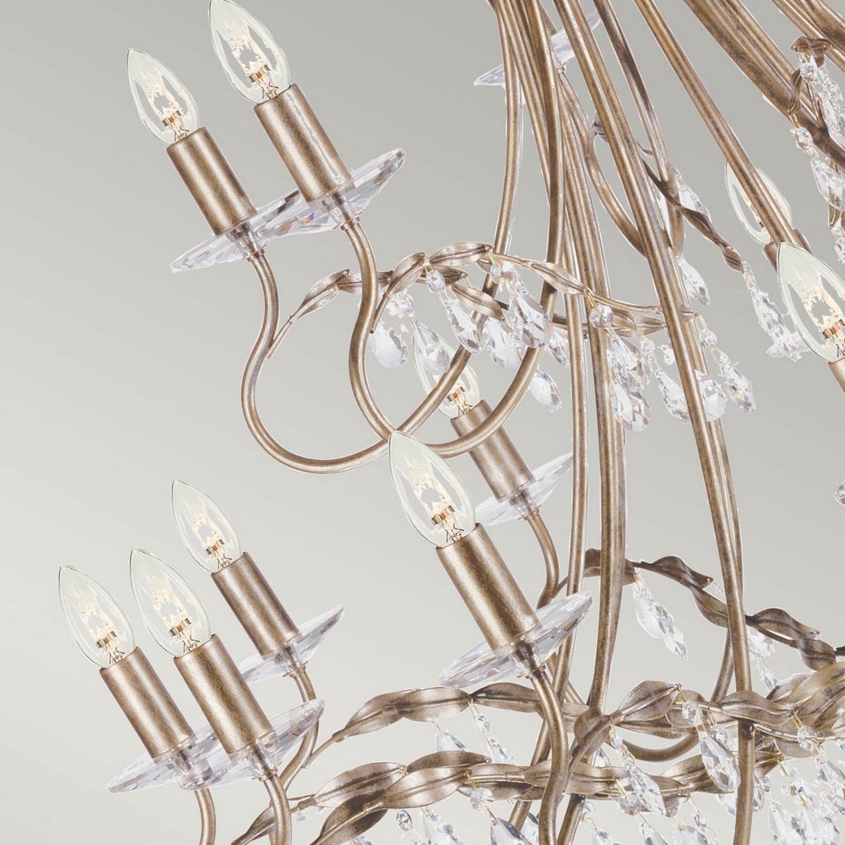 A close-up of the Christina 18 Light Chandelier, featuring elegant curved arms with crystal drops and small candle-shaped light bulbs. The chandelier's silver and gold patina shines beautifully against a soft gradient background, accentuating its intricate details.