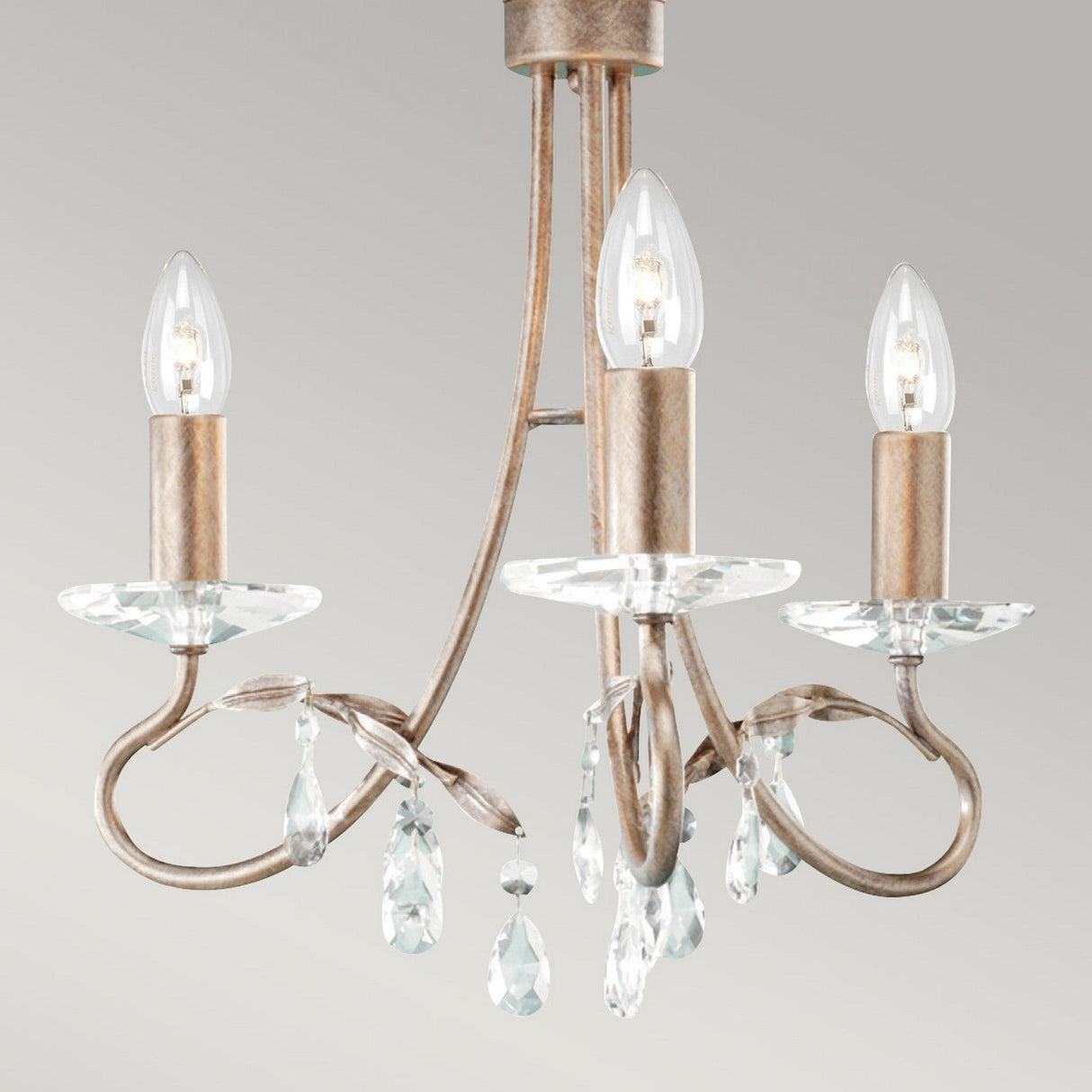 The Christina 3 Light Chandelier in Silver/Gold Patina features three arms adorned with a rustic metal finish and candle-shaped light bulbs. Each arm includes a glass plate and dangling crystal drops, set against a neutral background for an elegant touch to any room.