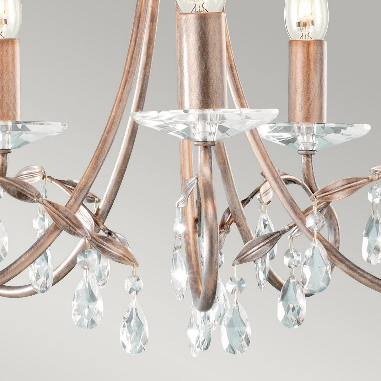 Close-up of the Christina 5 Light Chandelier in Silver/Gold Patina, featuring elegantly curved arms adorned with crystal drops. The crystals reflect light, creating a sparkling effect that enhances the chandelier's luxurious appearance. The background is a plain gray hue, highlighting its striking design.