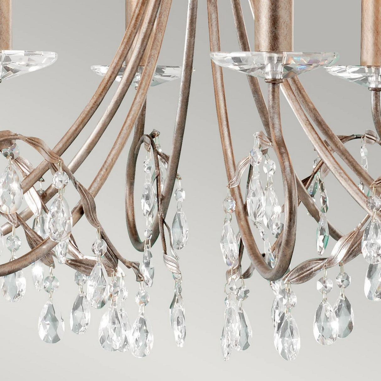 A close-up view of the Christina 8 Light Chandelier in Silver/Gold Patina showcases its ornate design, featuring curved metallic arms decorated with dangling prisms. The soft, neutral grey background highlights the chandelier's elegant design, making it an ideal choice for formal dining rooms.