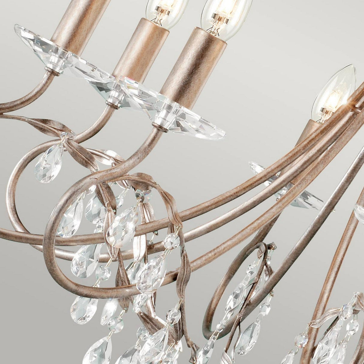 A close-up of the exquisite Christina 8 Light Chandelier, showcasing its sophisticated crystal design and intricately curling metal arms in a silver/gold patina finish with hanging droplets. Ideal for formal dining rooms, it features several candle-shaped light bulbs on top, casting a warm glow against a neutral backdrop.
