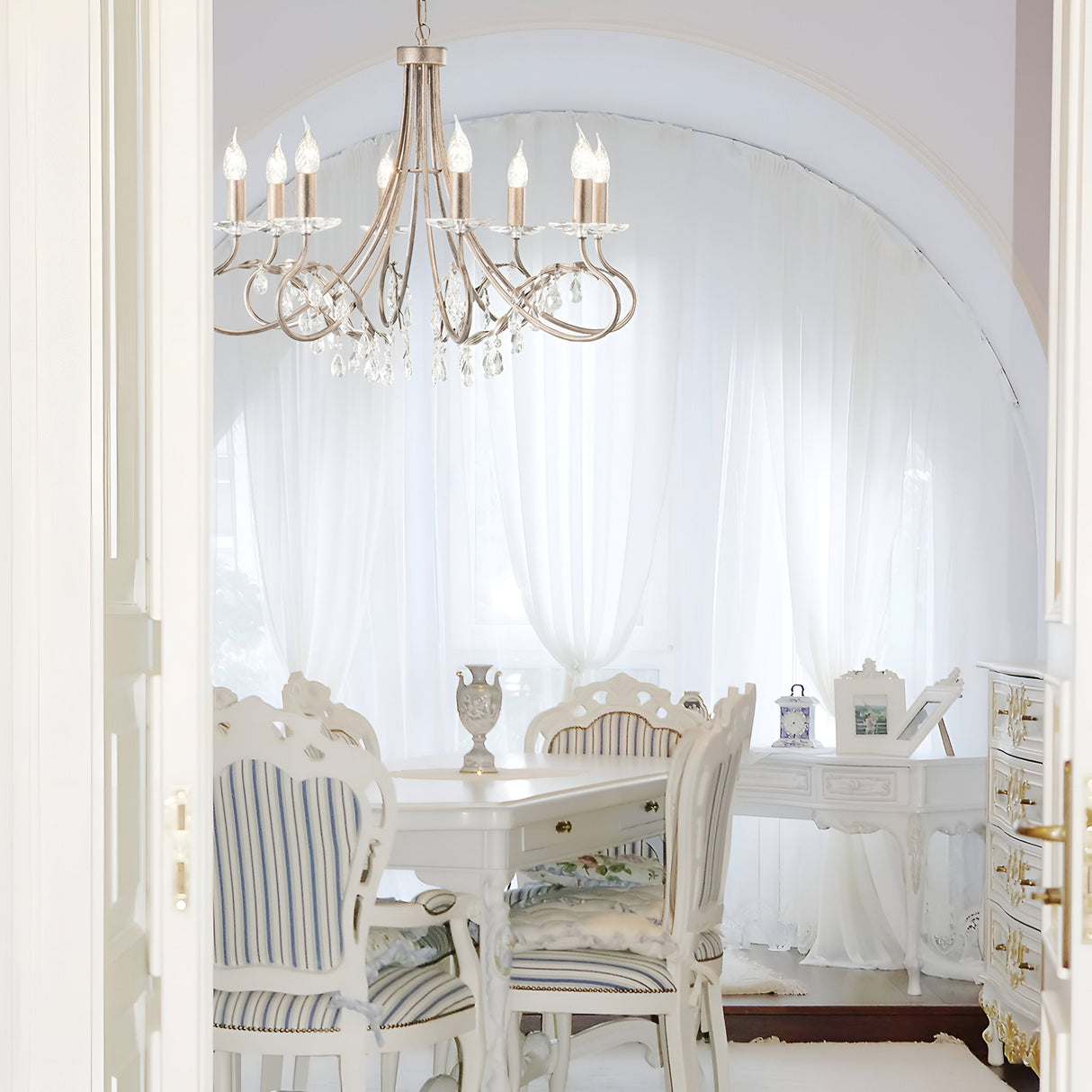 In a bright and elegant dining room, a white table is paired with striped chairs beneath the graceful Christina 8 Light Chandelier, adorned with crystal detailing and finished in Silver/Gold Patina. The space is further enhanced by sheer white curtains framing an arched window, creating an airy atmosphere.