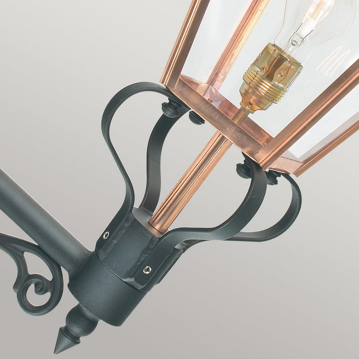 Presenting a detailed view of the Chelsea Outdoor 1 Light Wall Lantern - Copper, this elegant piece boasts intricate scrollwork on its arm. Crafted with a black metal frame and transparent glass panels, it beautifully displays the visible light bulb inside, making it an ideal choice for refined outdoor lighting.