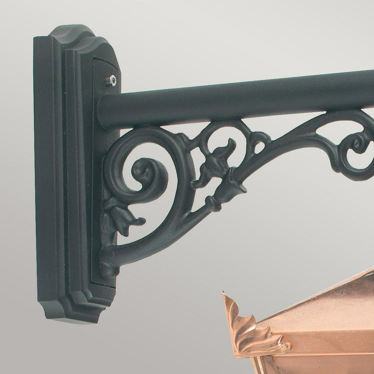 A close-up of the Chelsea Outdoor 1 Light Down Wall Lantern in copper features an ornate, black metal bracket with an intricate scrollwork design, supporting the lantern's weathered look. The wall mount is visible, and this fixture enhances exterior decor lighting with its timeless charm.