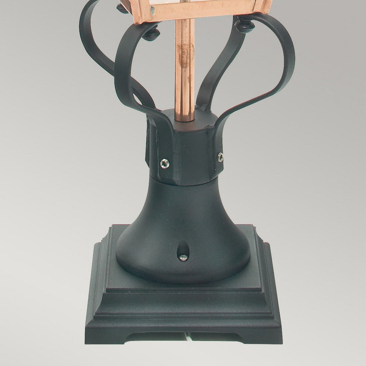 A close-up of the Chelsea 1 Light Pedestal Lantern in copper showcases its black decorative lamp base with a square shape and intricate metal detailing, reminiscent of a classic pedestal lantern. The central pole-like structure combines art and function, making it an ideal choice for outdoor decor against a plain gray backdrop.