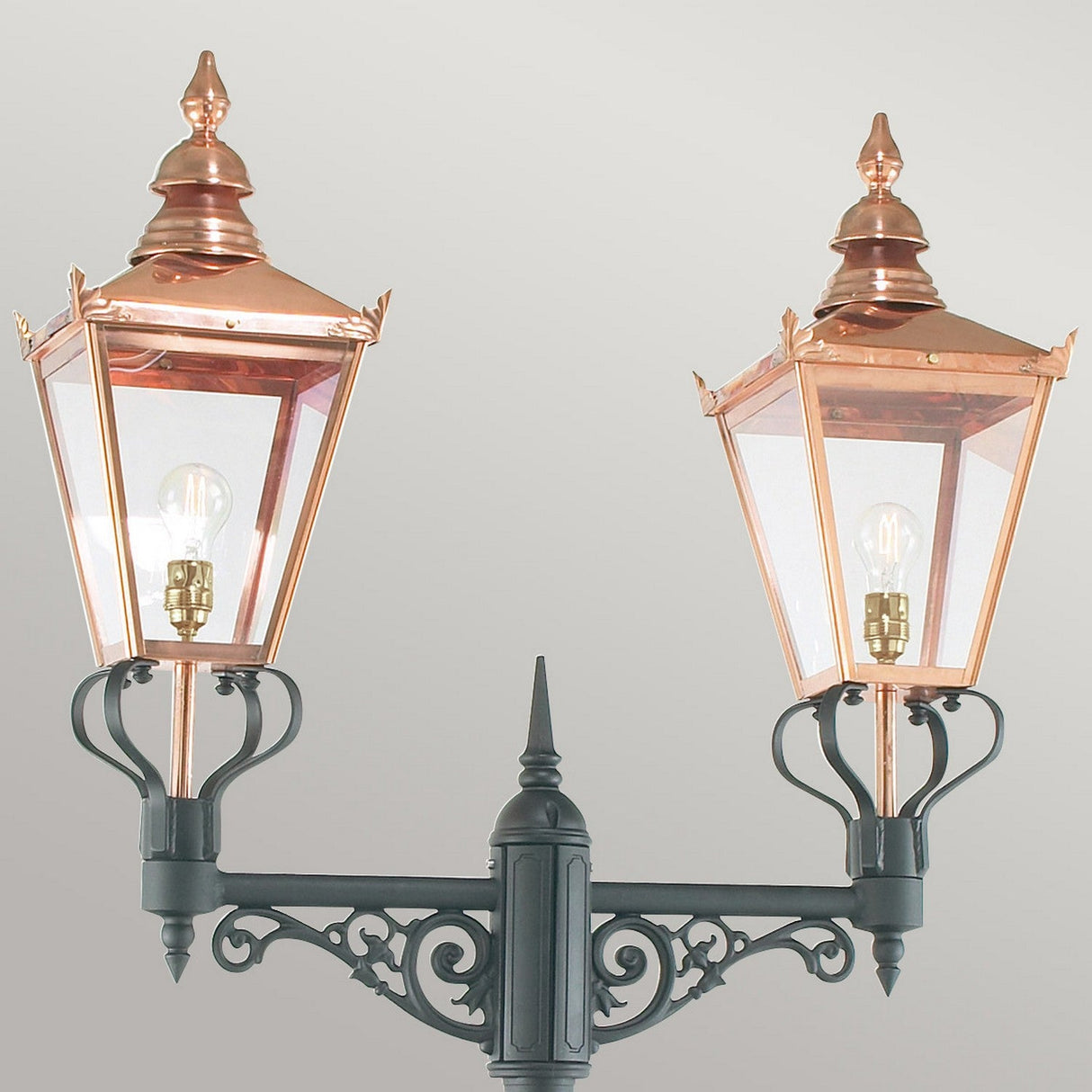 The Chelsea Twin Head Lamp Post - Copper & Black boasts two elegant copper lanterns with clear glass panels, beautifully positioned on a decorative black metal post. This outdoor lighting fixture highlights pointed tops and visible bulbs, creating a timeless copper and black aesthetic against a neutral backdrop.