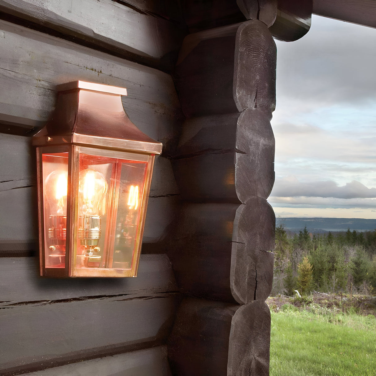 A Chelsea Outdoor 1 Light Small Half Lantern in copper, adorned with Edison bulbs, is mounted on a wooden log cabin. This handcrafted lantern enhances the charm of the forest and cloudy sky at dawn or dusk with its illumination. Its design harmonizes with nature while offering dependable IP44-rated lighting.