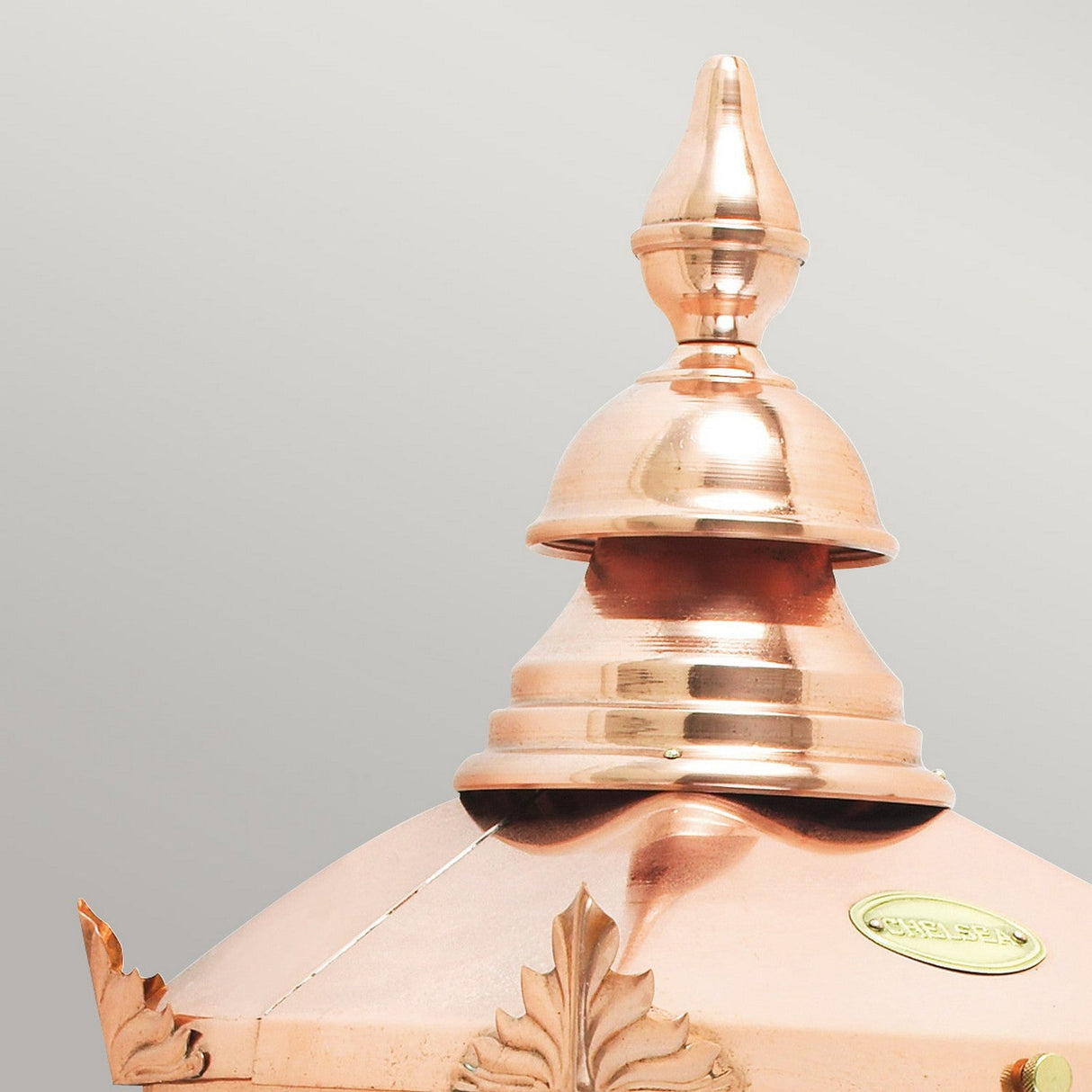 A close-up of the top portion of the Chelsea Outdoor 1 Light Half Lantern - Copper showcases its polished finish with decorative details and a pointed design. The smooth, shiny copper surface reflects the light beautifully, set against a plain background.