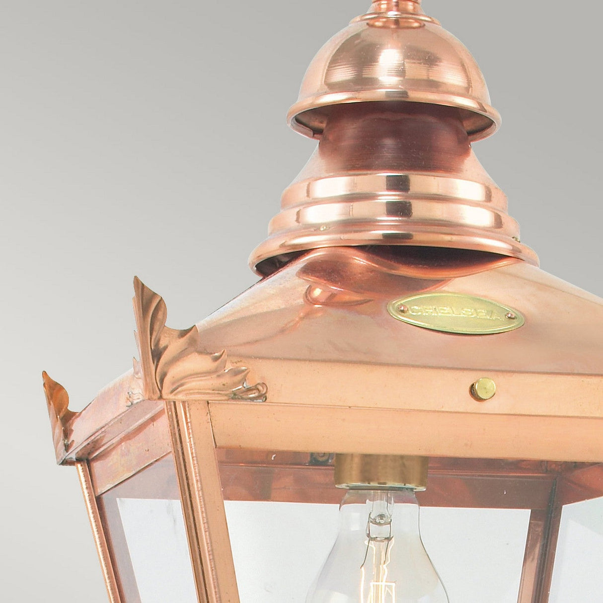 A close-up of the Chelsea Outdoor 1 Light Chain Pendant - Copper showcases a pendant with a clear glass panel, decorative edges, and a visible bulb inside. The fixture is mounted with an elegantly inscribed small round brass plate.