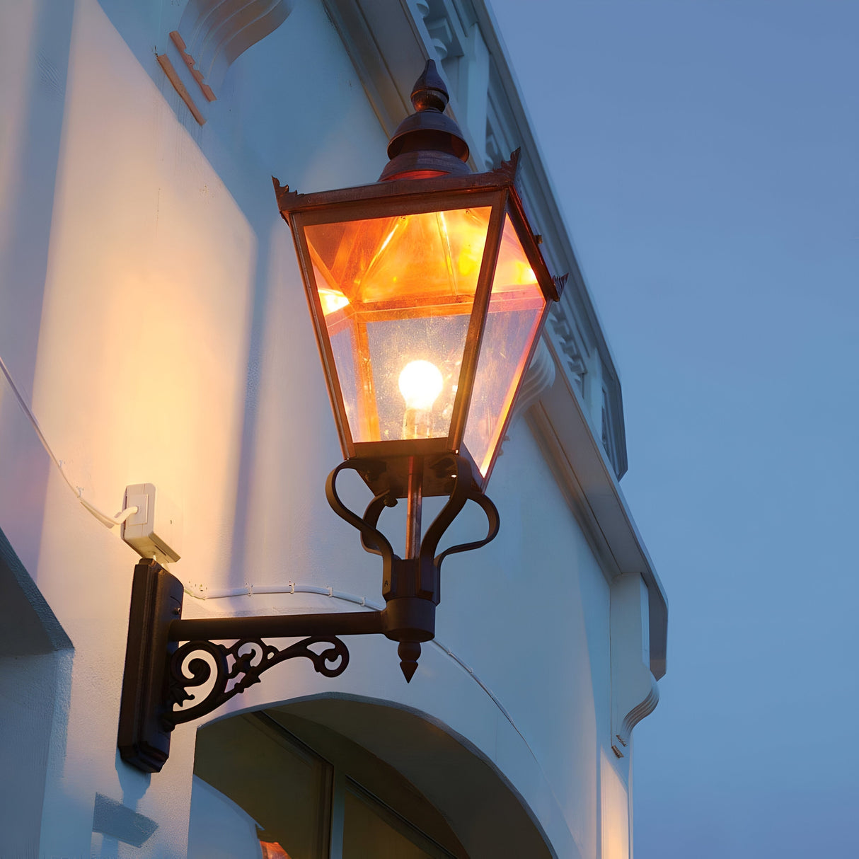 A Chelsea Outdoor 1 Light Grande Up Wall Lantern - Copper is elegantly mounted on the corner of a building, emitting a warm glow from its vintage-style design. The copper lantern illuminates the white facade with a cozy warmth, contrasting beautifully against the evening sky while showcasing its IP44 rating for durability.