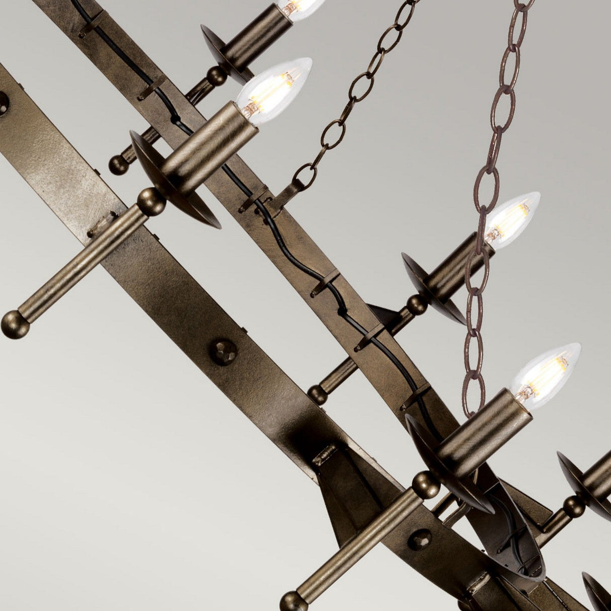 A close-up of the Cromwell 12 Light Chandelier - Old Bronze reveals its handcrafted solid iron structure adorned with multiple candle-shaped LED bulbs. The circular design is elegantly supported by chains and features an antique bronze finish, radiating a medieval charm that enhances the vintage appeal of any decor.