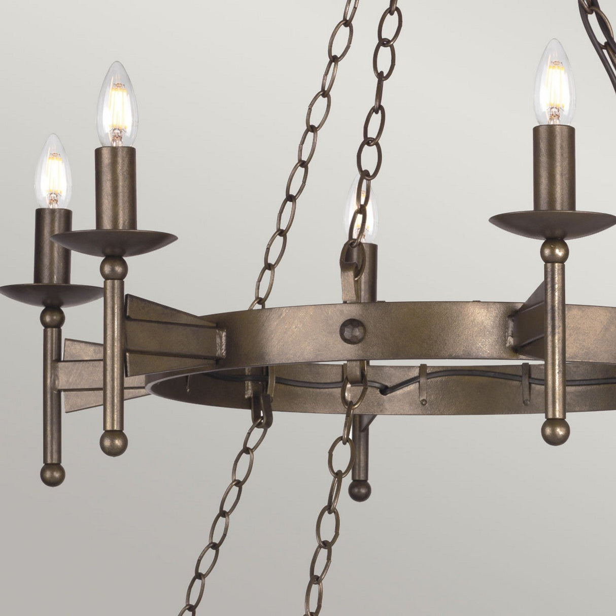 The Cromwell 18 Light Chandelier - Old Bronze showcases a medieval design with its rustic circular structure, featuring four candle-shaped bulbs suspended by chains from the ceiling. Its Old Bronze finish enhances the fixture's vintage appeal against a plain backdrop, evoking an air of timeless elegance.