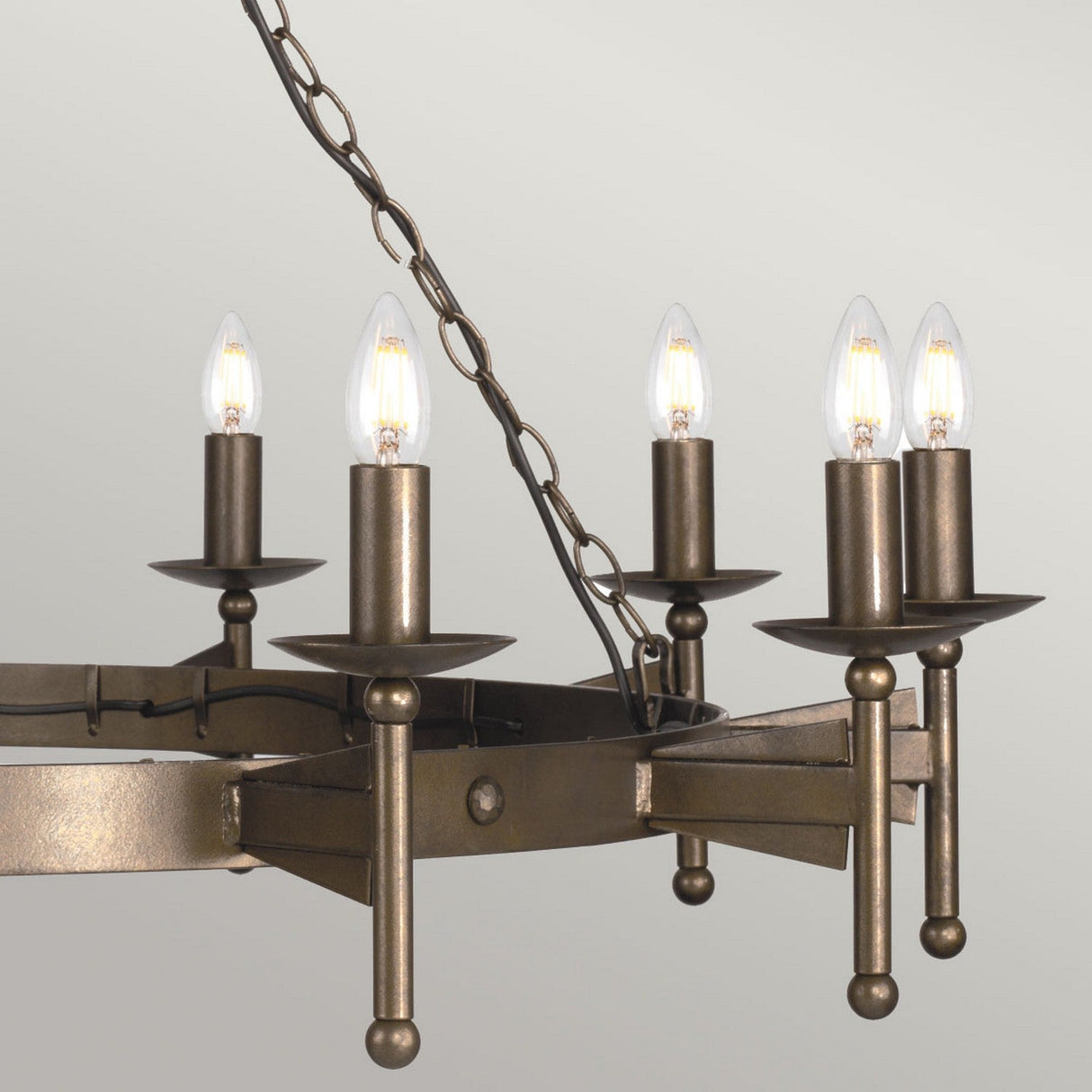The Cromwell 18 Light Chandelier in Old Bronze showcases a medieval design with its rustic appeal, featuring six candle-like light bulbs on a bronze metal frame. It hangs elegantly from a chain, with the old bronze finish creating a striking contrast against the light gray background.