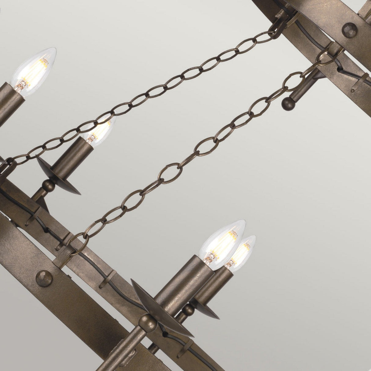 Close-up of the Cromwell 18 Light Chandelier in an Old Bronze finish. This fixture, with its candle-shaped light bulbs and chain links, embodies a medieval design reminiscent of antique charm. The background is a plain, light gray.