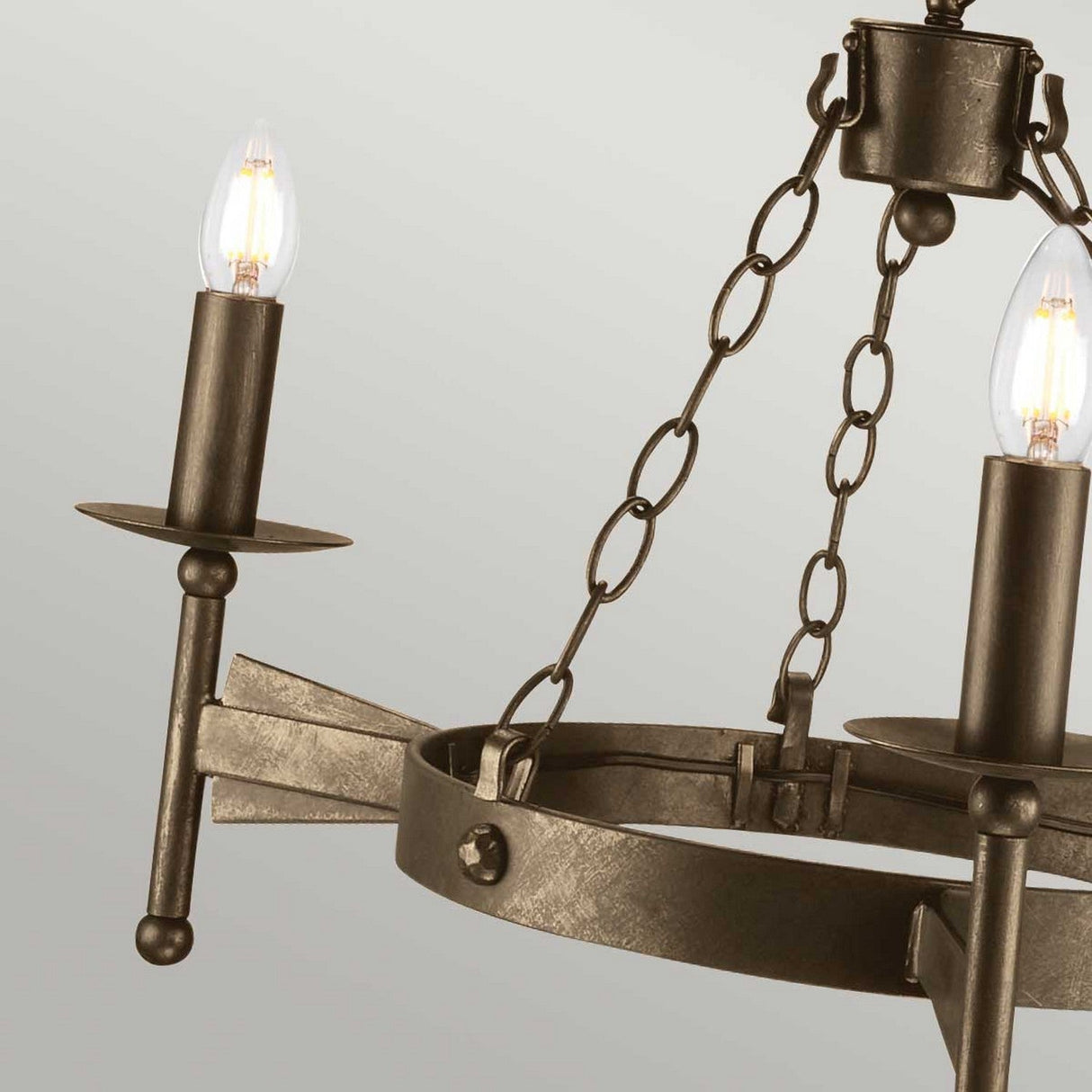 A close-up of the Cromwell 3 Light Chandelier showcases its rustic appeal with an old bronze finish and medieval design. It features two candle-shaped light bulbs, decorative chains, and a weathered look that beautifully complements the neutral background.
