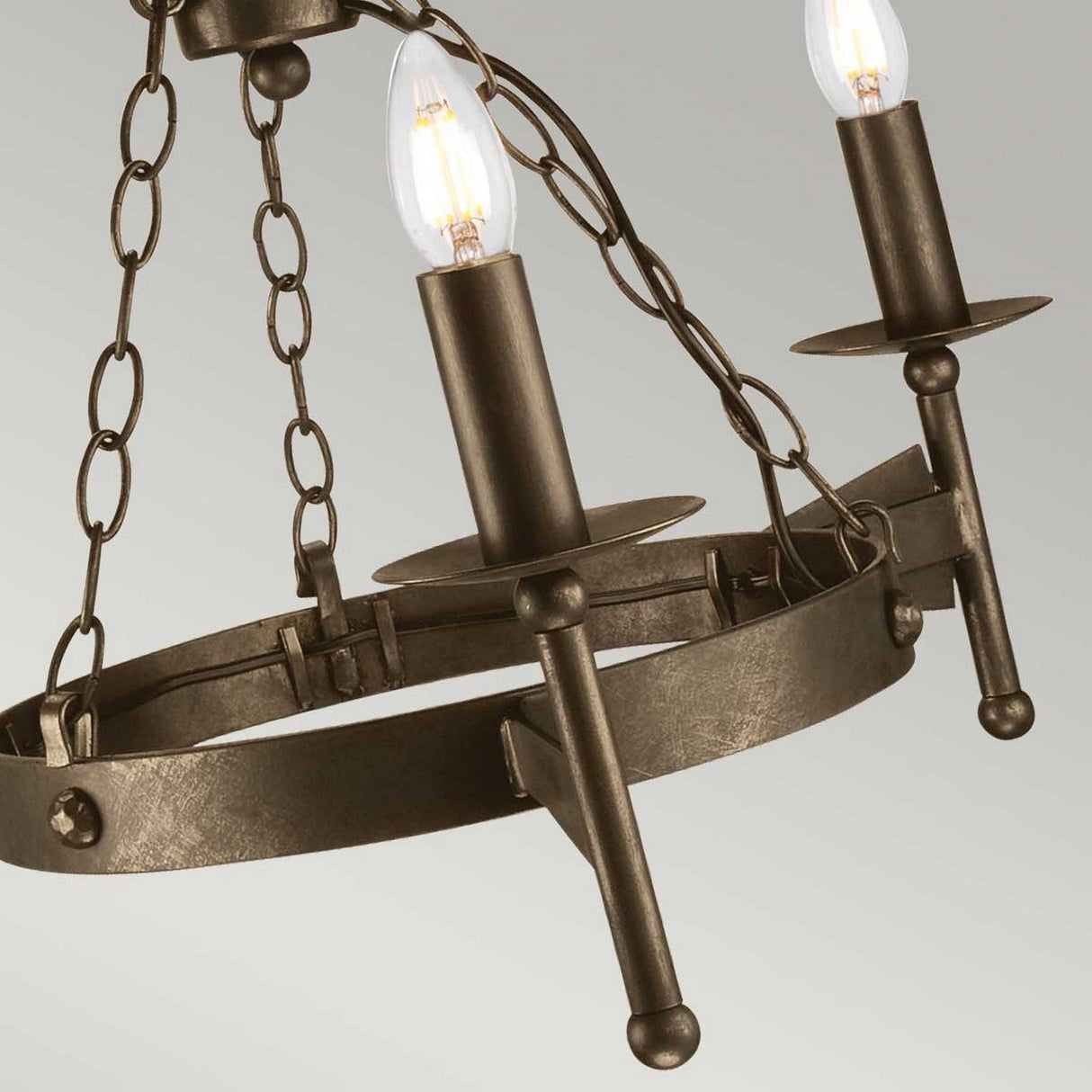 The Cromwell 3 Light Chandelier - Old Bronze features a rustic metal frame adorned with candle-like light bulbs. Its medieval design is suspended by chains, enhancing its vintage appearance, while the old bronze finish on the metal adds to its timeless allure.