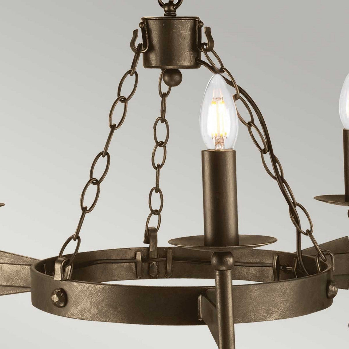 A detailed view of the Cromwell 3 Light Chandelier in Old Bronze reveals its exposed, candle-shaped bulbs. The chandelier's design, echoing medieval aesthetics, is characterized by its metal chains and circular frame, lending it an antique appearance against a simple light gray background.