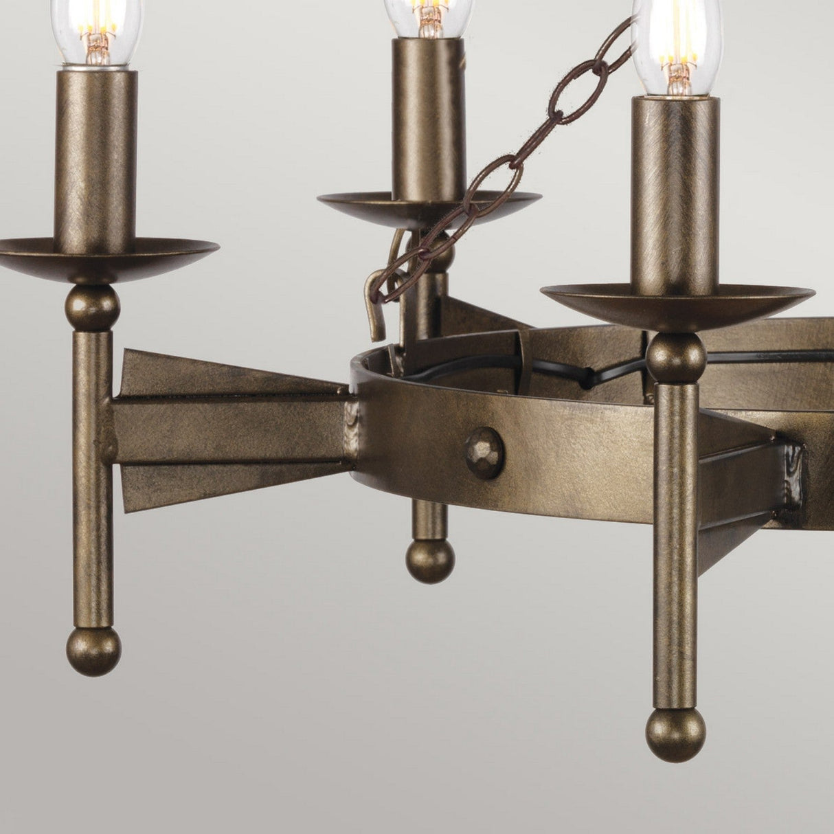 A close-up of the Cromwell 6 Light Chandelier in old bronze showcases a rustic design with three candle-style lights. Each light rests on simple cylindrical holders that are connected by chains to the central fixture. The unlit chandelier reveals intricate bulb details, echoing its elegance.