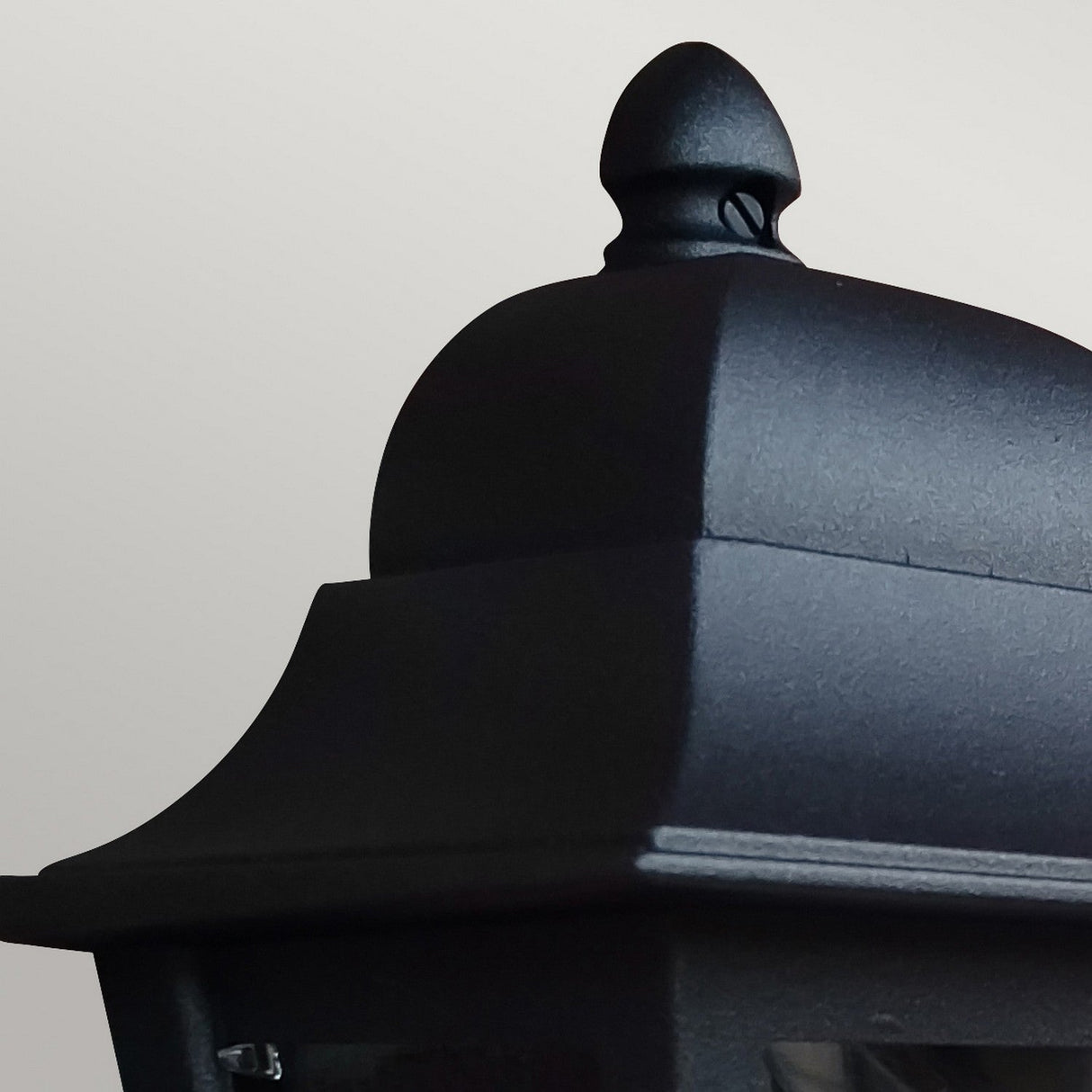 A close-up of the top section of the Deco Outdoor Wall Lantern - Black against a neutral background. The lantern showcases a smooth, matte finish with a rounded finial on top and a gently curved overhang.