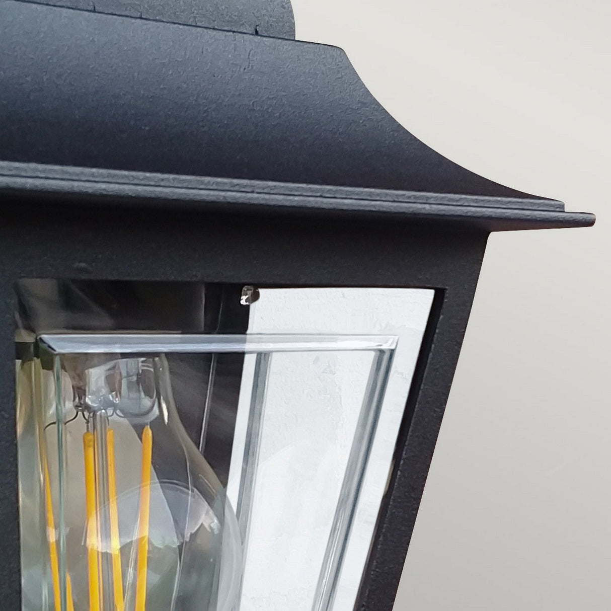 Close-up of the Deco Outdoor Wall Lantern - Black, featuring a clear glass panel that reveals an exposed filament bulb inside. Set against a plain, light gray background, this durable lantern in a sleek black finish enhances any exterior setting with elegance.