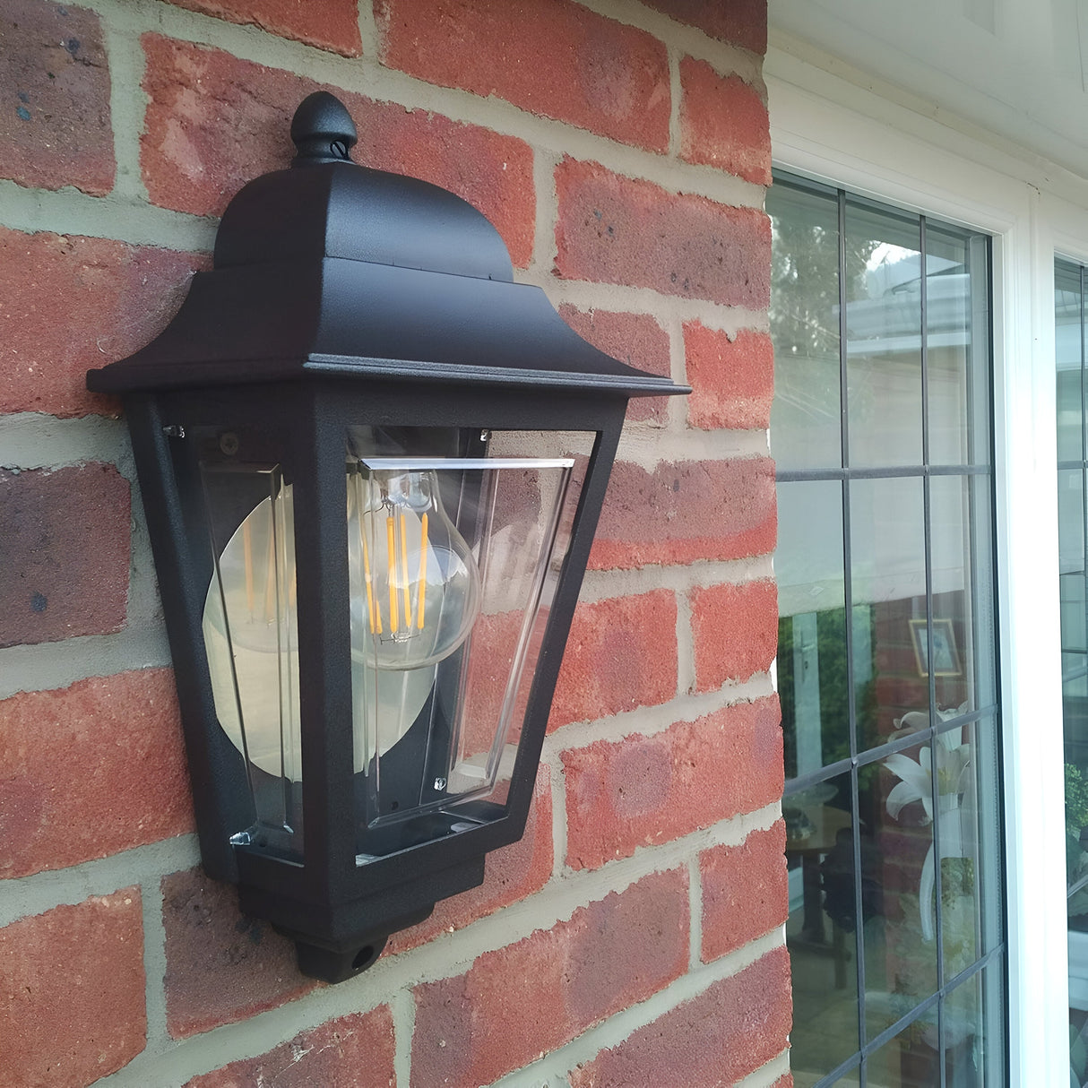 A Deco Outdoor Wall Lantern - Black, featuring a durable black finish, clear glass frame, and visible filament bulb, is mounted on a red brick wall beside a window reflecting the surrounding greenery.