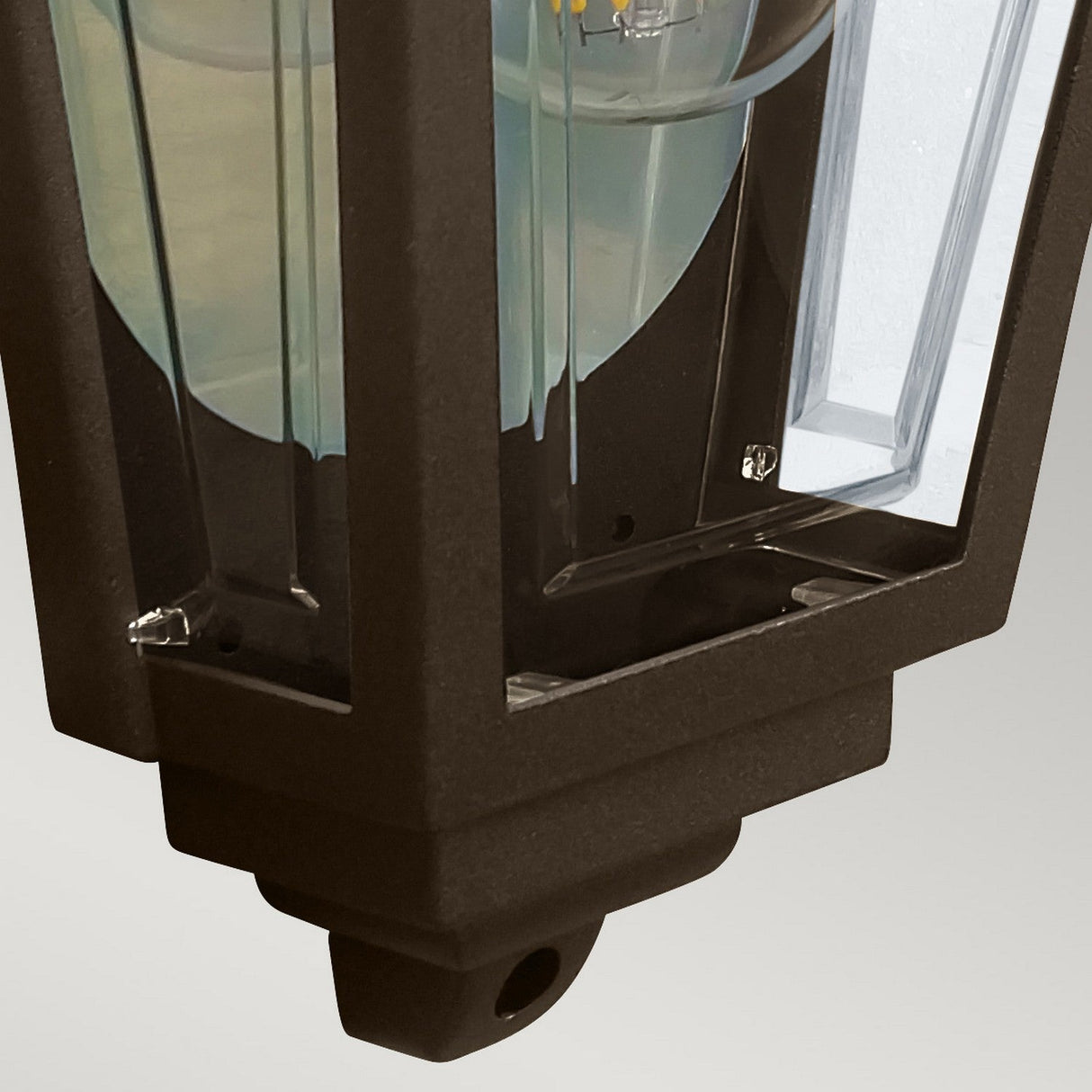 A close-up of the Deco Outdoor Wall Lantern - Bronze, showcasing a black-framed, lantern-style design with clear glass panels and a visible bulb. This fixture blends a traditional half-lantern style with modern, angular elements, set against a light gray background.