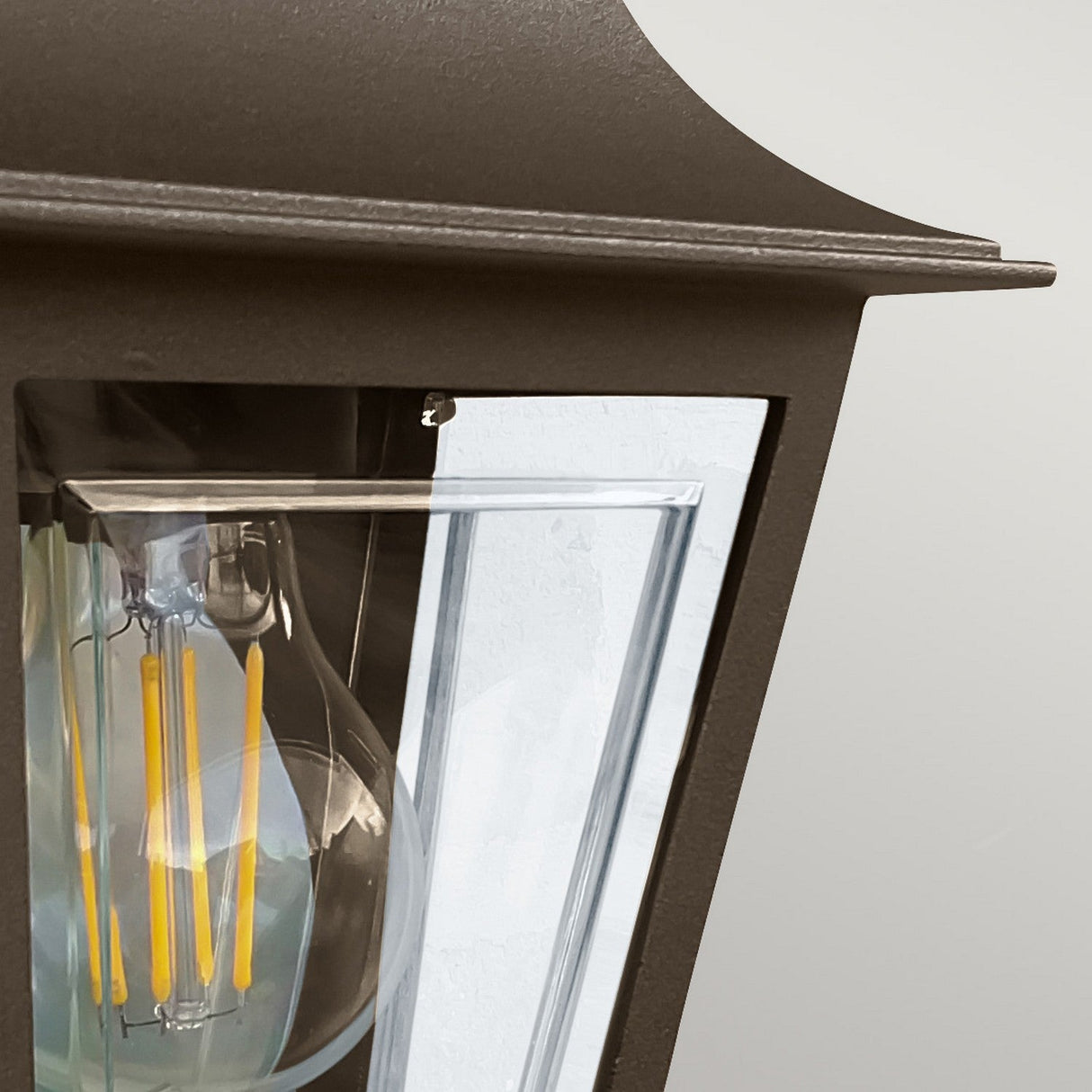 A close-up of the Deco Outdoor Wall Lantern in bronze, featuring clear glass panels that highlight an LED filament bulb. This lamp combines a modern, minimalist design with a gently curved top and hints of traditional half-lantern styling.