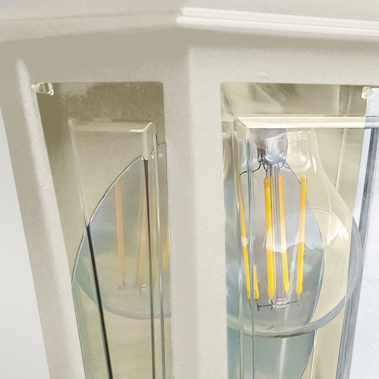 Close-up of the Deco Outdoor Wall Lantern - Cream, showcasing its vintage-style design with a cream finish and clear glass enclosure. It houses an LED filament bulb that emits a warm, ambient glow, accentuating the metal frame and reflective surfaces, making it an ideal outdoor wall light.