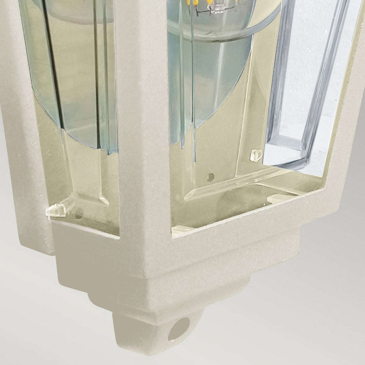 A close-up of the Deco Outdoor Wall Lantern - Cream, showcasing its geometric design with a transparent cubic frame made from durable aluminum, featuring internal reflective surfaces and a visible bulb element, set against a plain light gray background.