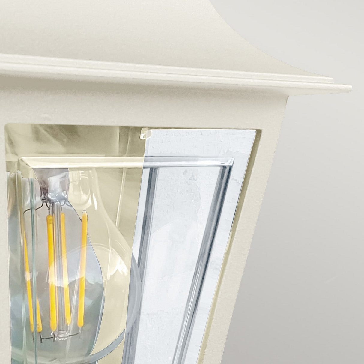 Close-up of the Deco Outdoor Wall Lantern with a visible clear glass bulb and yellow filaments. It features a modern lantern design with a cream finish and sleek glass panels, enhancing its contemporary appeal.