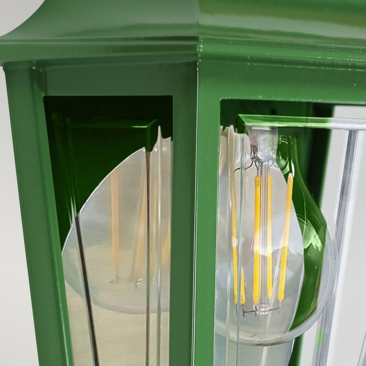 A detailed view of the Deco Outdoor Wall Lantern - Green showcases its aluminum structure with a green finish and transparent glass panels revealing an LED bulb. This outdoor wall light is distinguished by its reflective surfaces and sleek metal craftsmanship, set against a simple backdrop.