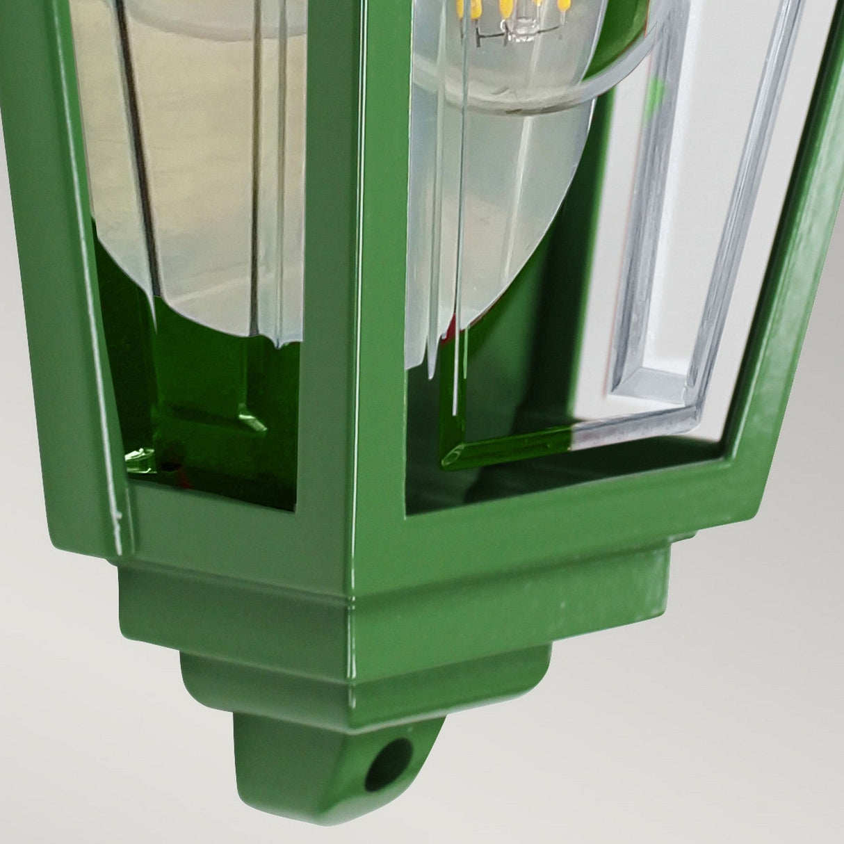 The Deco Outdoor Wall Lantern - Green features a detailed close-up view showcasing its clear glass panels that elegantly expose the bulb inside. Hanging gracefully from its top, the lantern's metal frame glistens with a shiny finish, making it an ideal choice for adding decorative lighting to your outdoor space.