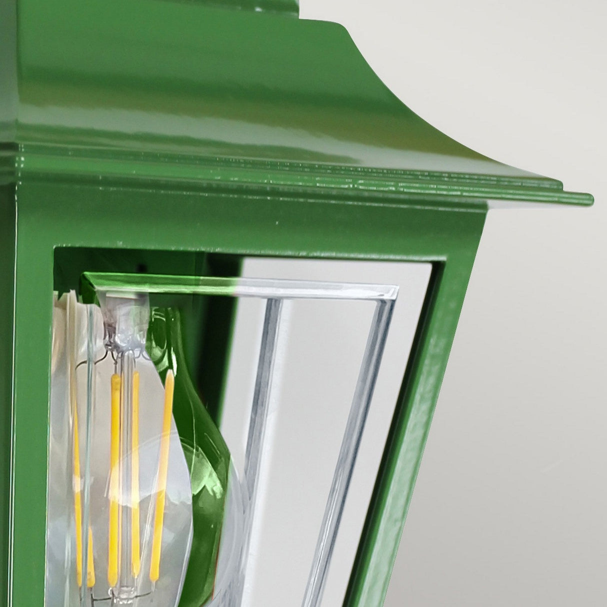 Close-up of the Deco Outdoor Wall Lantern - Green, showcasing its elegant green finish. The glass panels reveal a softly glowing candle-style LED bulb inside, and the fixture's slightly angled roof design adds a touch of sophistication to any setting.