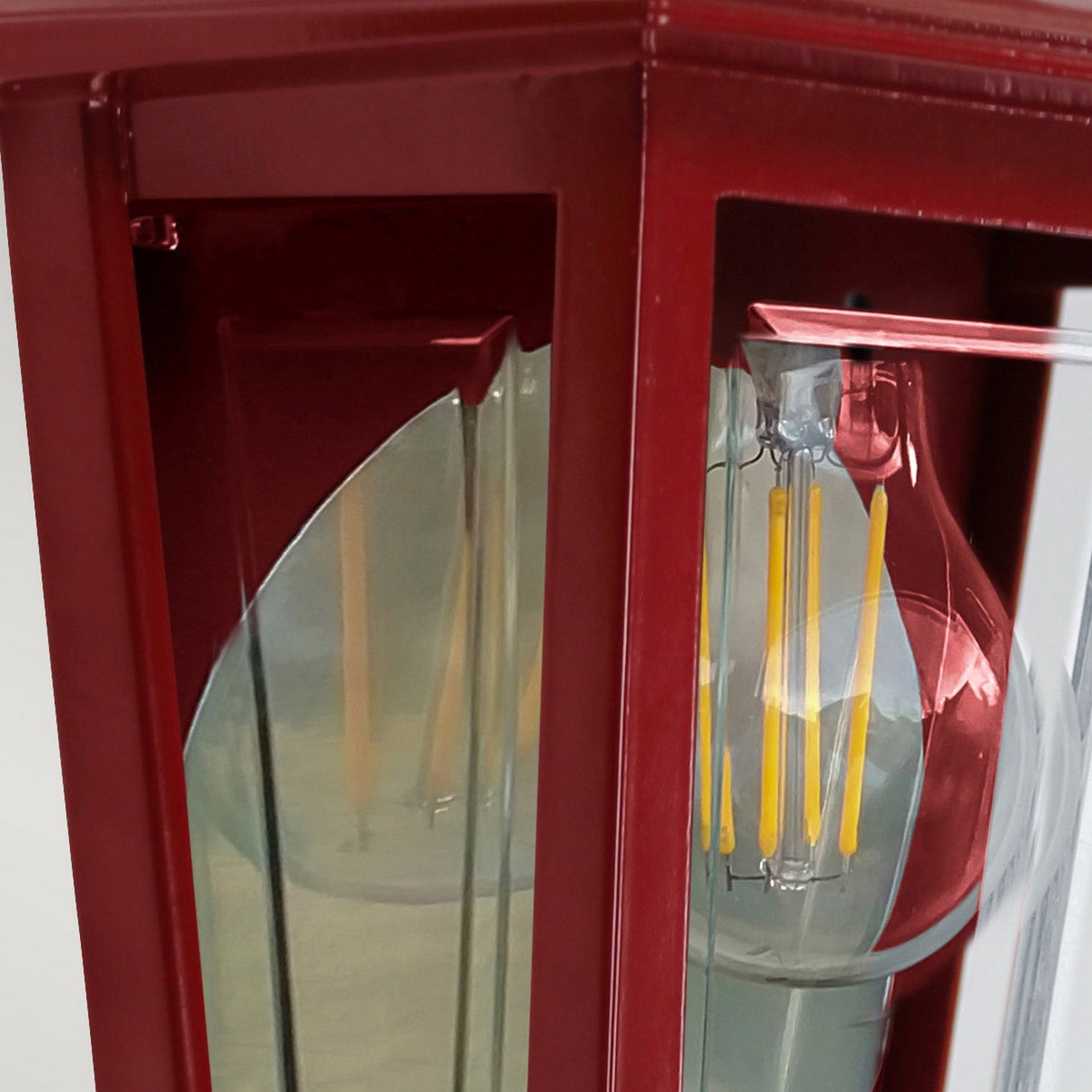 Close-up of the Deco Outdoor Wall Lantern - Red, displaying a red-framed box with a transparent front that highlights an LED filament bulb inside. The reflective glass and mirrored backplate accentuate the glowing yellow filaments of the bulb.